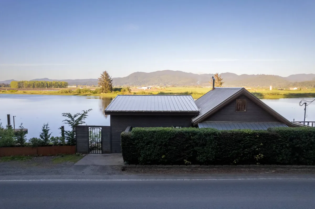 This exterior image features the Wild Run River House set against a scenic river and mountain backdrop. The house is shown nestled close to the water, surrounded by greenery, with ample outdoor space for guests and their pets to explore. The inviting setting hints at tranquil adventures along the riverbank, perfect for canine companions.