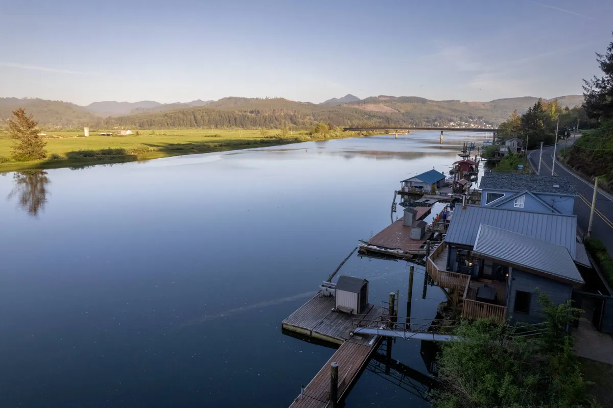 A serene view of a calm river with docks, houses on stilts, and a mountainous backdrop. The natural shoreline and tranquil setting create an inviting atmosphere for nature walks and playtime with pets along the water's edge.
