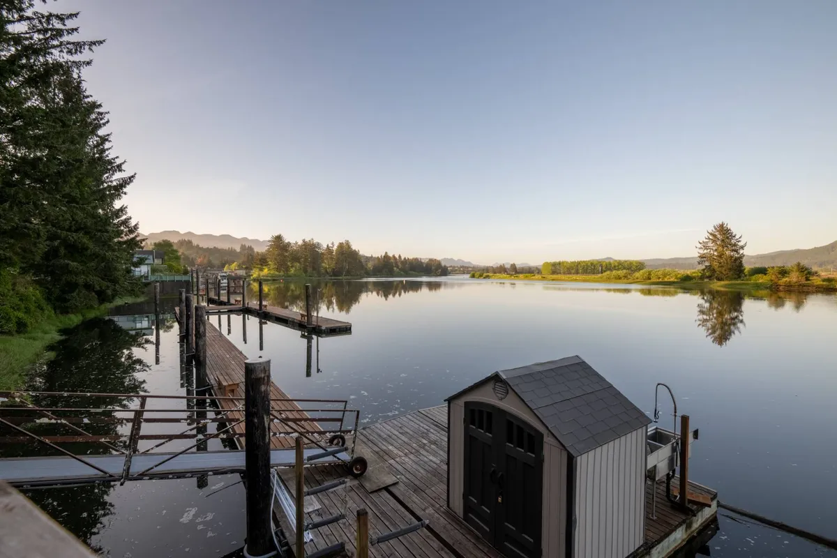 A calm water surface reflects the sky and trees, with a dock and a small shed visible along the shore. This peaceful riverside setting encapsulates the possibilities for fishing, boating, and tranquil moments on the water.