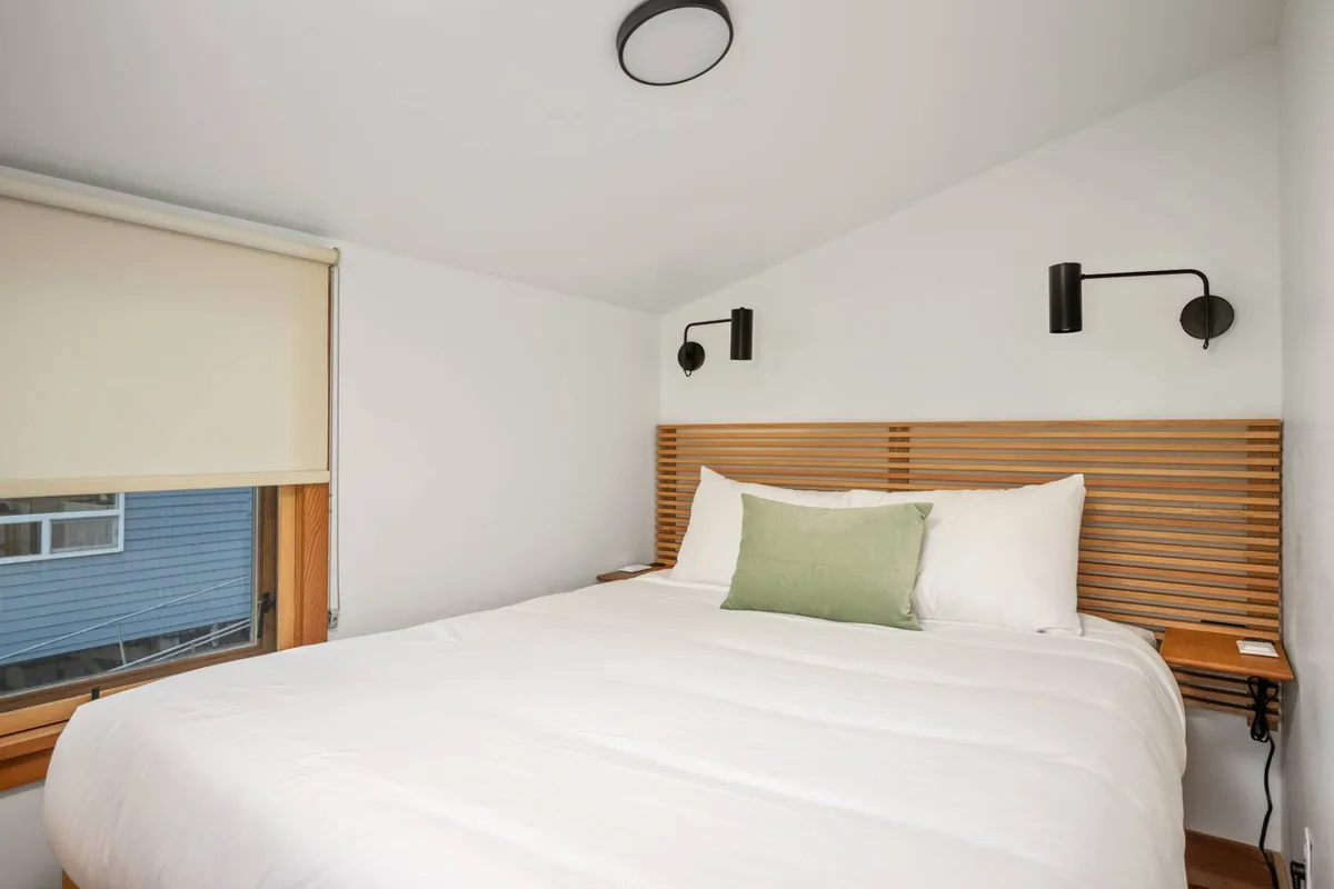 A tranquil bedroom scene featuring a queen bed with crisp white linens, a wooden headboard, a green accent pillow, and modern wall sconces. The room feels bright and peaceful, bathed in soft natural light.