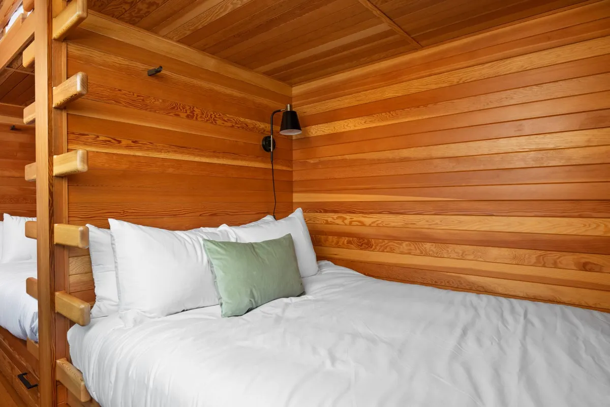 Intimate view of a wooden bunk bed with plush white bedding and a green accent pillow, set against a warm wood-paneled wall. The arrangement encourages shared, close quarters, implying togetherness.