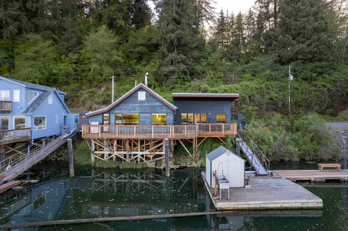 The image showcases a waterfront property with a wooden deck overlooking the river. There are docks extending into the water and trees surrounding the area, providing a shaded, natural environment ideal for relaxation and outdoor activities with dogs.