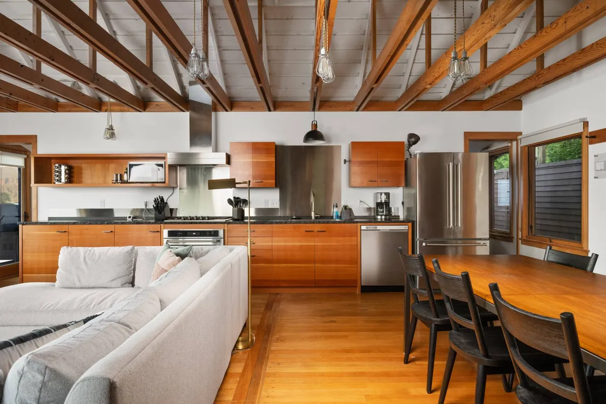 This image shows an inviting open-concept kitchen, dining, and living space. The kitchen island and dining table are in close proximity to a comfy sofa, all tied together by exposed wooden beams and ample natural light, suggesting an ideal setting for group activities like dining, board games, and morning coffee with a view.