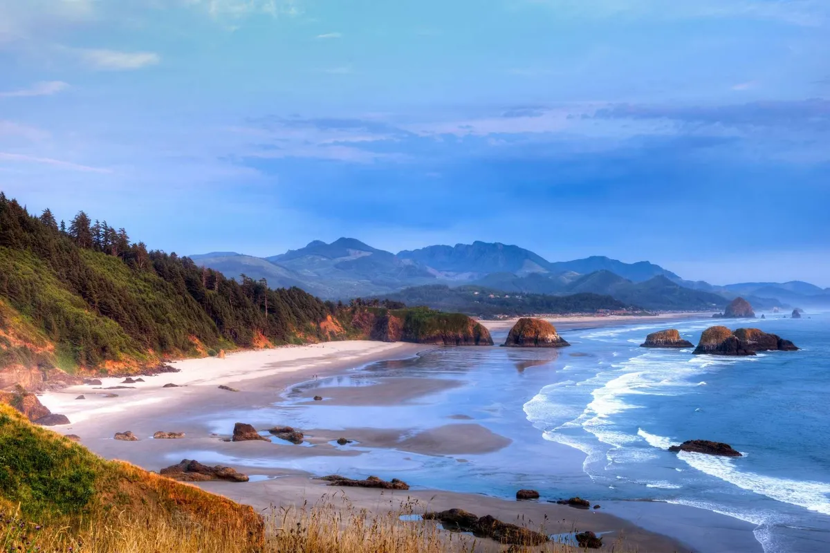 A tranquil sandy beach with rolling ocean waves, prominent rock formations, a distant mountain range, and a tree-lined shore. The gentle light suggests dusk, enhancing the peaceful coastal vibe.