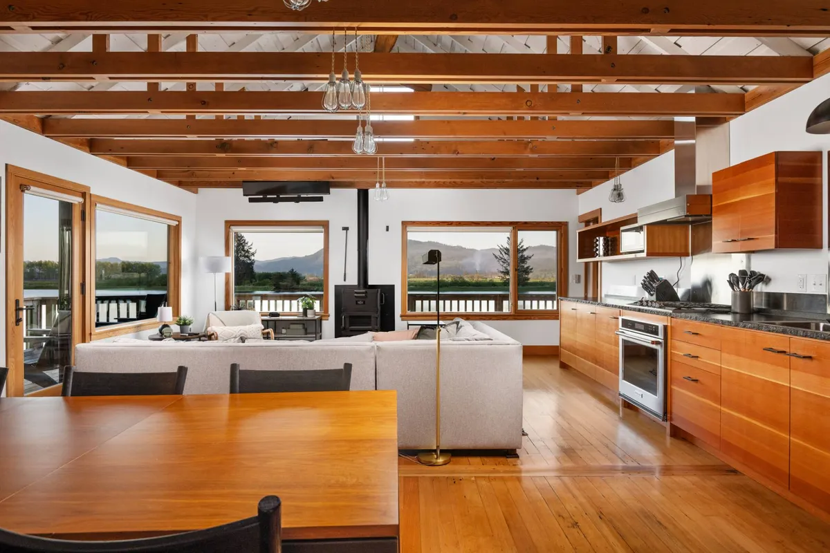 The image showcases a sunlit open-concept living space with expansive windows framing peaceful river and mountain views. A modern kitchen is fully equipped with sleek appliances, while a wooden dining table and a plush sectional sofa create inviting spaces for gathering and relaxation. Warm décor and natural light create a cozy, welcoming ambiance perfect for quality time with family or friends.