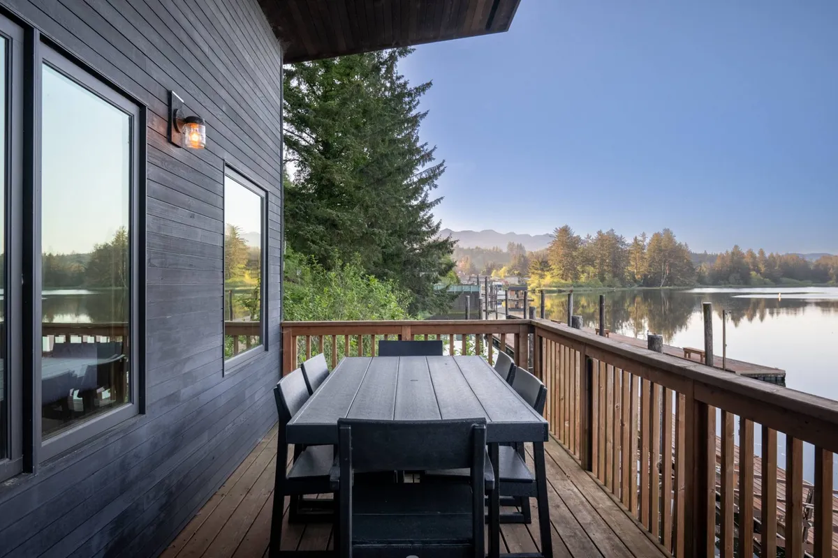 An inviting wooden dining table is set outdoors, framed by lush trees and offering a picturesque view of the nearby water and mountain backdrop. The scene suggests relaxed outdoor dining, socializing, and enjoying tranquil, natural surroundings—perfect for unwinding after a day exploring local flavors.