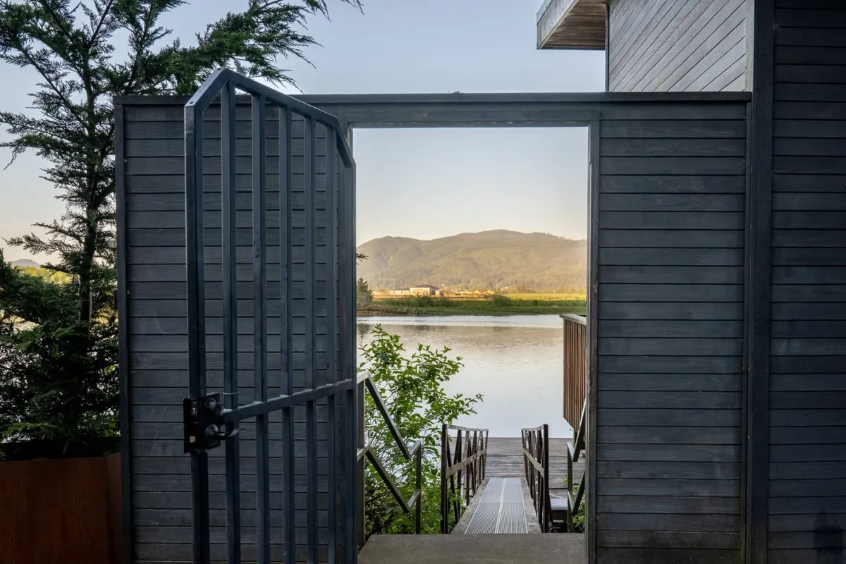 A tranquil exterior view shows an open gate leading down a lush, green path directly to the water, with a mountain backdrop. The scene evokes the ease of access to the river and the natural beauty that surrounds the property, inviting exploration and outdoor enjoyment.