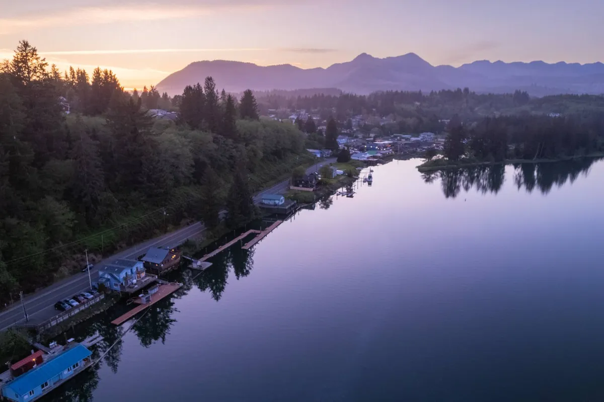 A wide view of a serene waterway bordered by lush forest, a mountain backdrop, and quaint houses along the shore. The scene hints at tranquil boating, fishing, and the easy access to local attractions by water, connecting directly to the section’s emphasis on arriving downtown Nehalem by paddle.