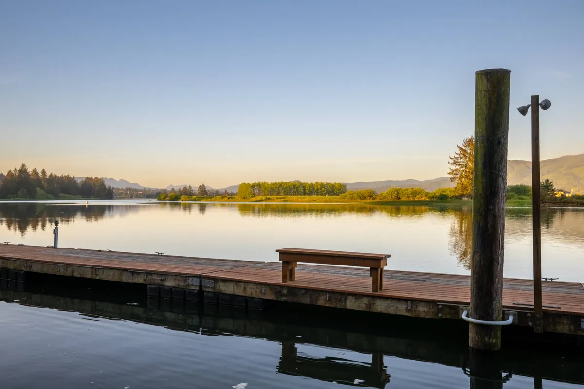 A peaceful dock extends over calm waters, with a bench inviting quiet moments of reflection. The surrounding landscape includes distant mountains and greenery, reinforcing the tranquil riverside atmosphere. The dock’s placement emphasizes a personal, immersive connection to the water and nature.