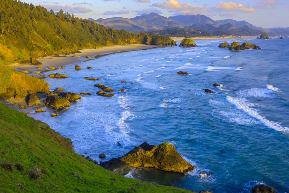 A sweeping view of the Pacific Ocean with a wide sandy beach, dramatic rock formations dotting the shore, a bordering forest, and distant mountains under a serene sky. The scene is vibrant yet peaceful, capturing the essence of the Oregon Coast's natural beauty.
